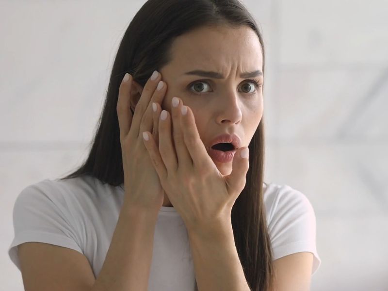 začuđena devojka dodiruje svoje lice i gleda se u ogledalo